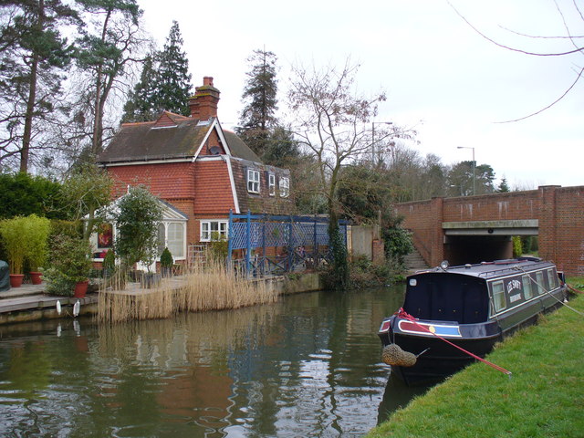 Red brick house by a canal with a narrowboat and bridge, surrounded by trees and plants on a cloudy day.