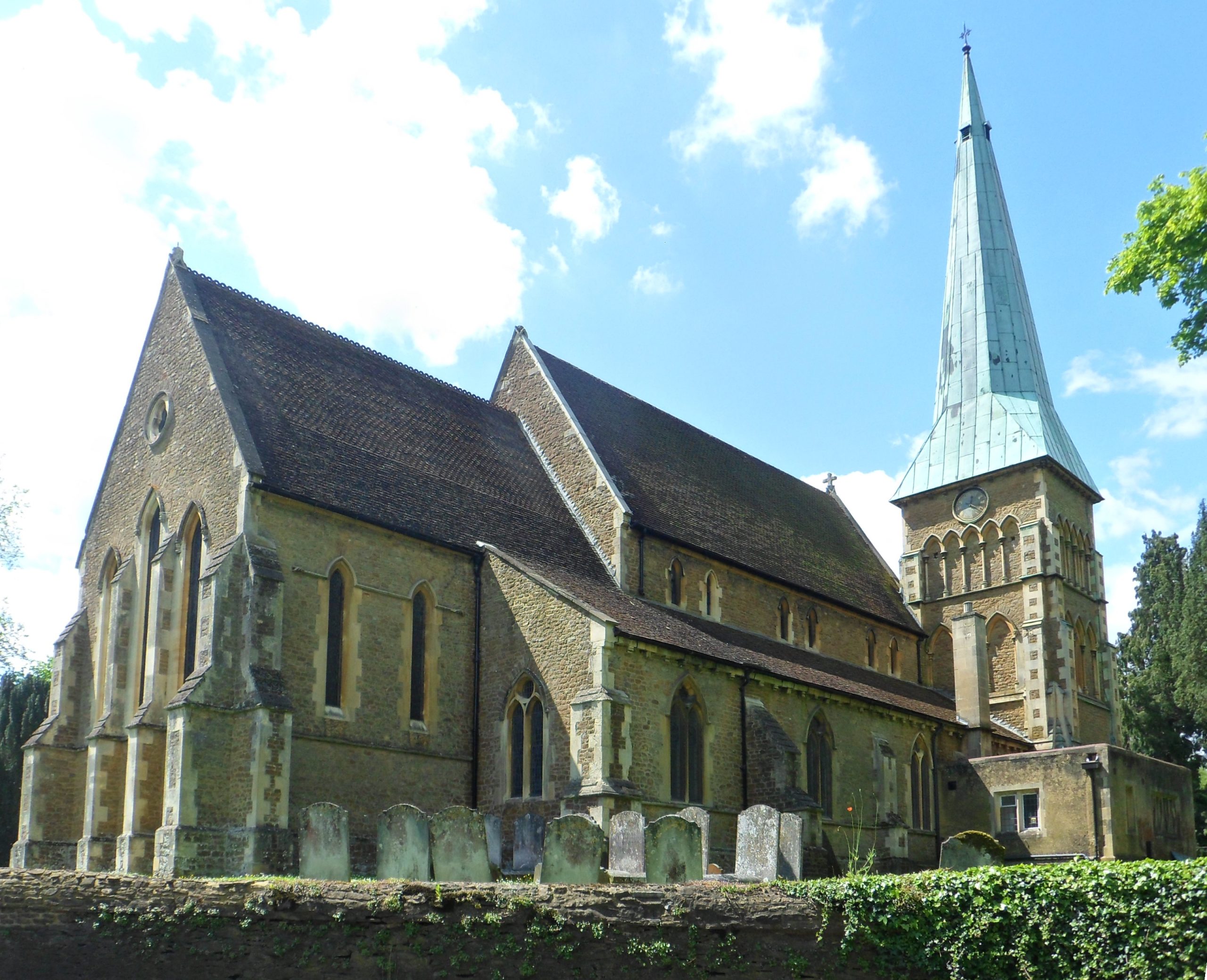 Historic stone church with tall spire, surrounded by gravestones, set against a bright blue sky and lush greenery.