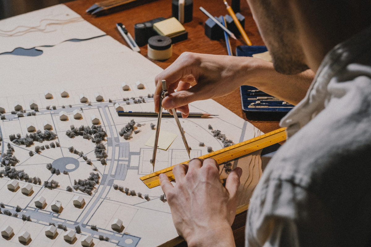 Architect measuring a scale model of a neighborhood using a compass and ruler on a desk filled with tools.
