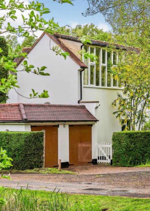 Charming countryside cottage with green hedges and trees, featuring white walls and a red-tiled roof.